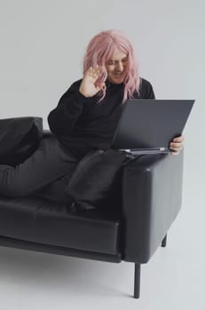 Portrait of a smiling man in a pink wig, sits on a sofa, communicates via video link on a laptop. White background. Vertical frame.