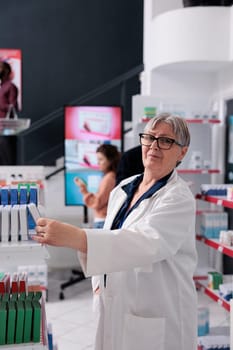 Senior pharmacist woman standing beside drugstore shelves arranging pills packages for help clients, worker offering medical service and support. Diverse people in pharmacy buying vitamins
