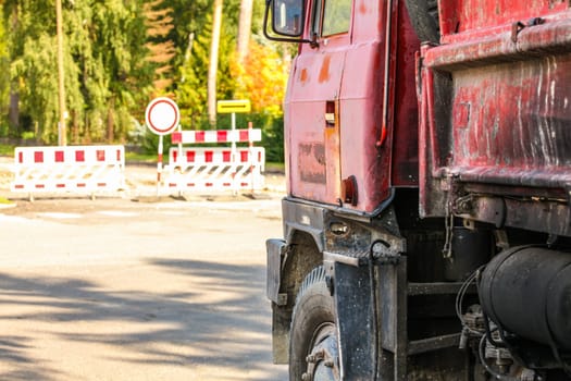 Closeup of old rusty and dirty red truck side, paint peeling on cargo area wall, large tires visible, blurred road block sign and closed path in background. Roadworks illustration.
