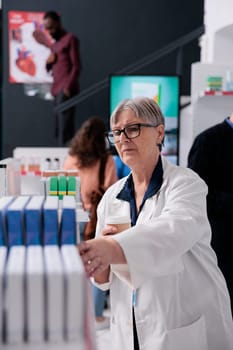 Elderly pharmacist in white coat looking at pills packages checking pharmacy shelves while helping people with health care treatment. Diverse customer buying vitamins, supplements, health care service