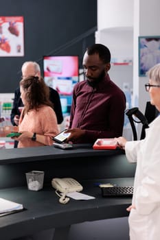 Drugstore client standing at pharmacy counter putting phone on pos terminal paying pills prescription with modern phone, buying vitamins and supplements. Health care service and support