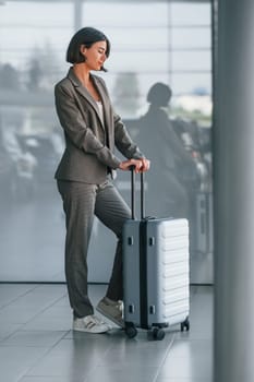 Against wall with reflection. Woman with luggage is standing indoors. Conception of tourism.