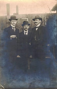 GERMANY - CIRCA 1905: Vintage cabinet card shows dandy men wear bowler hats, luxury garment and one holds cigar. Antique black and white photo. Photo taken in backyard