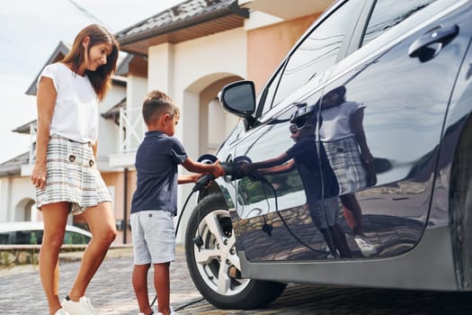 Charging the car. Woman with little boy near the the modern automobile at daytime.