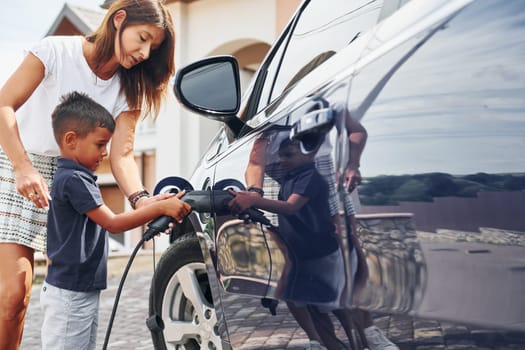 Charging the car. Woman with little boy near the the modern automobile at daytime.