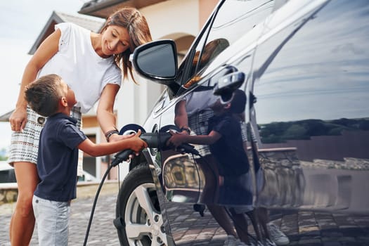 Charging the car. Woman with little boy near the the modern automobile at daytime.