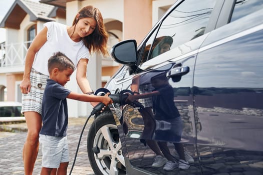 Charging the car. Woman with little boy near the the modern automobile at daytime.