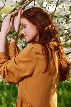 a happy, modest woman is standing in an orange dress near a flowering tree and holding her hands on the branches leans her head to them. High quality photo