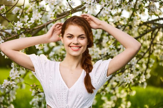 portrait of a joyful woman in a light dress against the background of a flowering tree, raising her hands to her head. High quality photo