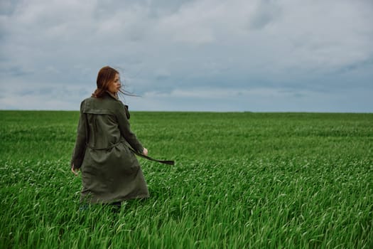 a woman in a long raincoat stands in a green field in windy weather with her back to the camera. High quality photo