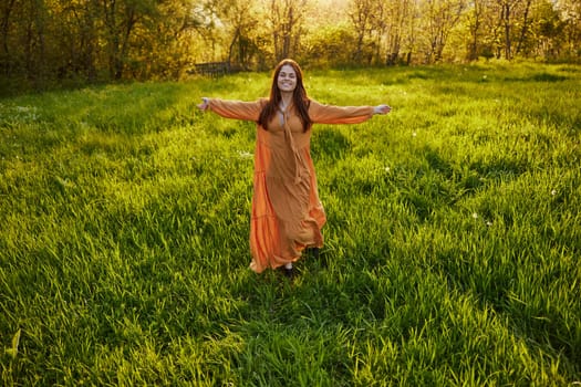 an attractive, slender, red-haired woman stands in a wide, green field during sunset in a long orange dress enjoying unity with nature and relaxation, raising her arms to the sides while facing the camera. High quality photo