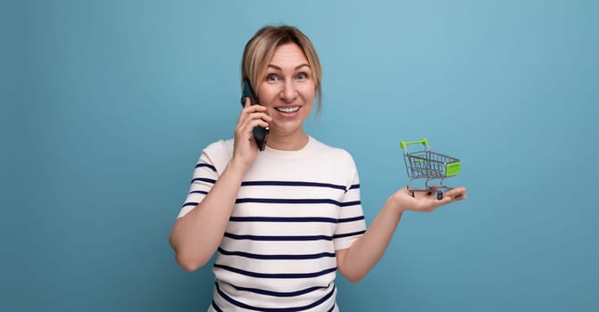 horizontal photo for banner of attractive shopper girl talking on phone holding shopping cart on blue background with copy space.
