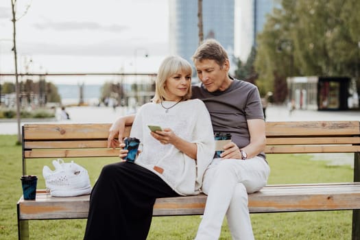 An adult mature happy couple in love sitting on bench with phone outdoors in park. A blonde caucasian man and woman spend time together and drinking coffee. Senior wife and husband walking outside
