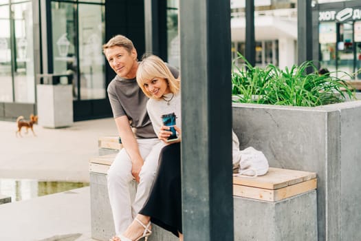 An adult mature happy couple in love sitting on bench outdoors in city street park. A blonde caucasian man and woman spend time together and drinking coffee. Senior wife and husband walking outside