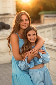 Portrait of mother and daughter in blue dresses with flowing long hair against the backdrop of sunset. The woman hugs and presses the girl to her. They are looking at the camera
