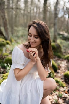 Portrait of a woman in the forest. She is sitting in a white dress on a meadow with snowdrops in a spring forest.