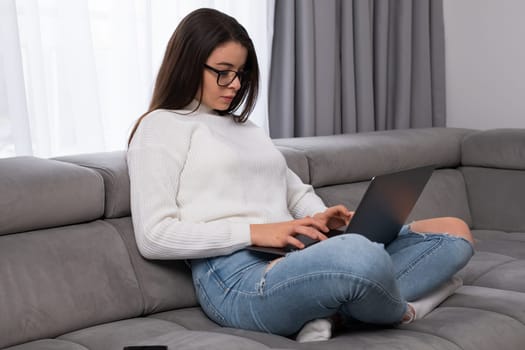 Serious woman in eyeglasses using laptop checking email sitting on sofa or study online.
