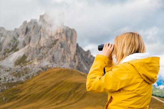 Young blonde woman looks in binoculars to high mountain pass in the Dolomites, the Giau Pass.