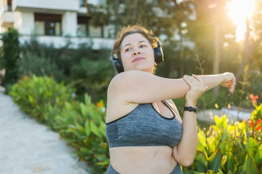 Adorable fat woman in tracksuit is engaged in fitness outdoor side view portrait. Young overweight woman lunges outdoors on warm summer day. Healthy lifestyle and weight loss.