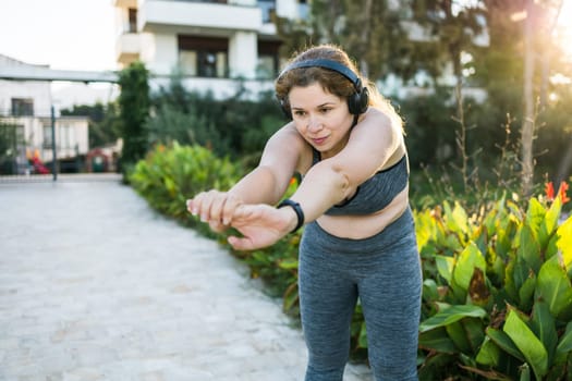 Adorable fat woman in tracksuit is engaged in fitness outdoor side view portrait. Young overweight woman lunges outdoors on warm summer day. Healthy lifestyle and weight loss.