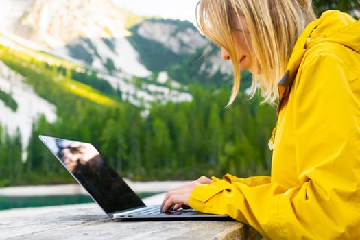 The girl is focused on working at a computer in nature with a beautiful view of the mountains and the lake. Freelancer works remotely from the office in sunny day.