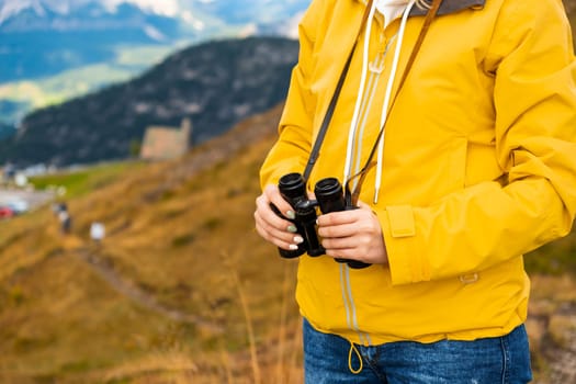 Close up a woman in a yellow jacket holding binoculars with mountains on the backgtound.