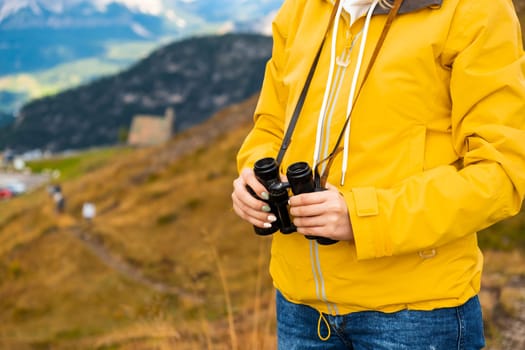 Close up a woman in a yellow jacket holding binoculars with mountains on the backgtound.