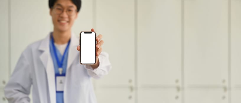 Doctor wearing white uniform showing smartphone with blank screen for advertisement. Telehealth online applications concept.