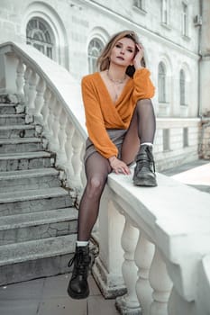 A middle-aged woman looks like a good blonde with curly beautiful hair and makeup on the background of the building. She is wearing a yellow sweater