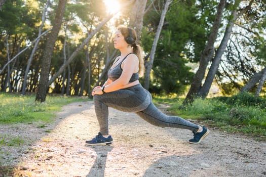 Adorable fat woman in tracksuit is engaged in fitness outdoor side view portrait. Young overweight woman lunges outdoors on warm summer day. Healthy lifestyle and weight loss.