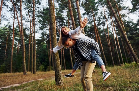 Woman is riding her man. Happy couple is outdoors in the forest at daytime.