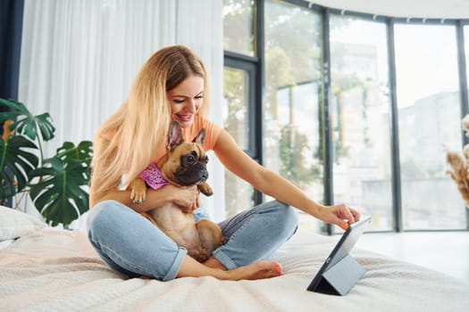 With modern tablet on bed. Woman with pug dog is at home at daytime.