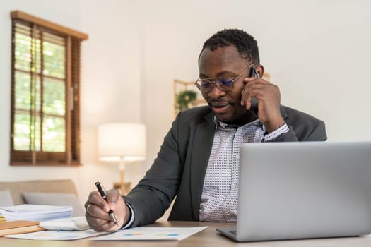 Portrait of handsome African black young business man working on laptop and talking phone at office desk...