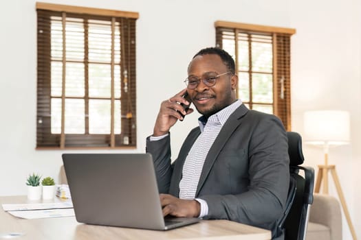 Portrait of handsome African black young business man working on laptop and talking phone at office desk...