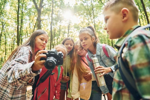 Holding camera. Kids in green forest at summer daytime together.