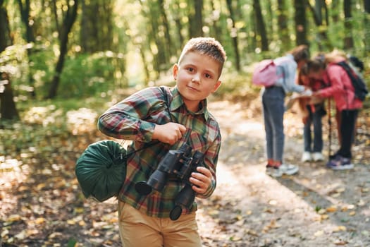 Boy with binoculars standing in front of his friends. Kids in green forest at summer daytime together.