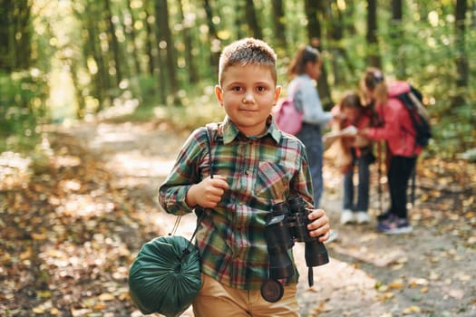 Boy with binoculars standing in front of his friends. Kids in green forest at summer daytime together.