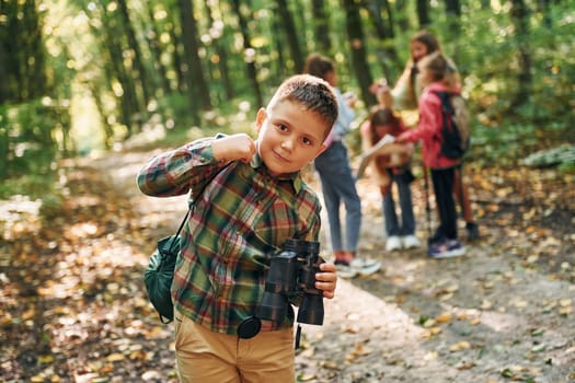 Boy with binoculars standing in front of his friends. Kids in green forest at summer daytime together.