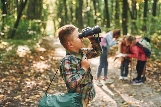 Boy with binoculars standing in front of his friends. Kids in green forest at summer daytime together.