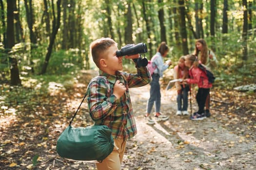Boy with binoculars standing in front of his friends. Kids in green forest at summer daytime together.