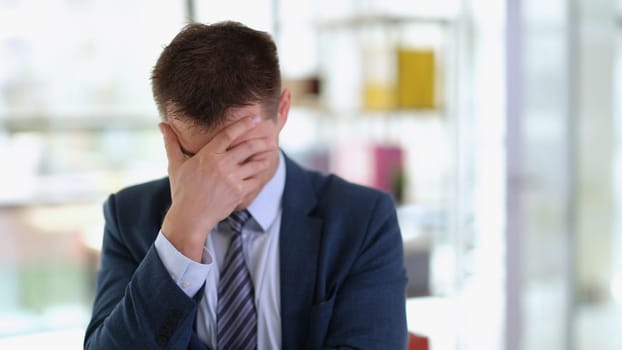 Sad businessman in suit covering face with his hand in office. Problems and difficulties of doing business concept