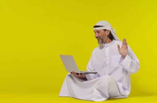 Concept online communication. Muslim person sitting on floor greets someone with their palm raised while holding a laptop in their hand. Copy space allows for easy customization and versatility and global connection through technology. High quality photo