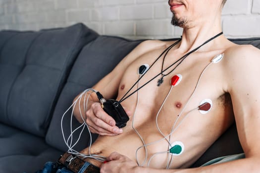 a man sitting on a sofa with a Holter heart monitor connected. Holter Monitor. electrocardiogram. study of the work of the heart, cardiology. Health control. Holter monitor. Medical diagnostics. Health care, hospital. ECG sensors. . Treatment heart diseases.