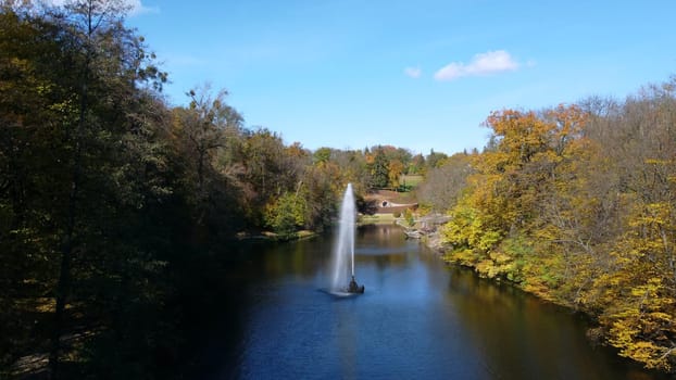 Fountain with rainbow in middle of lake between trees with yellow leaves in park on sunny autumn day. Decorative fountain in center of lake. Natural park arboretum in autumn. Natural landscape. Aerial