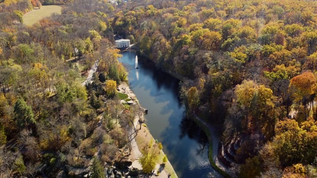 Beautiful panoramic autumn landscape park, many trees with yellow leaves, lake with fountain in center, architecture, big stones, paths walkways, people walking on sunny autumn day. aerial drone view