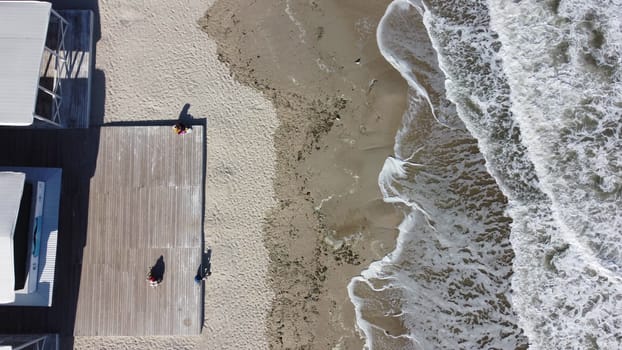 People sitting on a sandy beach near the sea and sea waves with white foam. People rest and relaxing near the sea. Flying over a sandy beach with people on a sunny day. Top view. Aerial drone view