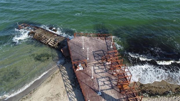 Old wooden pier with people resting and old concrete breakwater on the sandy beach of the sea shore on a sunny day. Public beach. Sea coast, sea shore. Coastal seascape. Top view. Aerial drone view