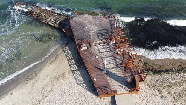 Old wooden pier with people resting and old concrete breakwater on the sandy beach of the sea shore on a sunny day. Public beach. Sea coast, sea shore. Coastal seascape. Top view. Aerial drone view