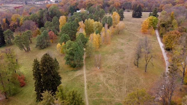 Panoramic view of large natural park with trees with yellow green red leaves,meadow, architecture and people walking along dirt park paths on autumn day. Beautiful nature background.