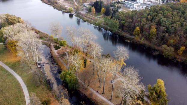 Flying over autumn park. Many trees with yellow green and fallen leaves, lakes, river, people walking along dirt paths in park on autumn day. Top view. Aerial drone view. Beautiful natural background